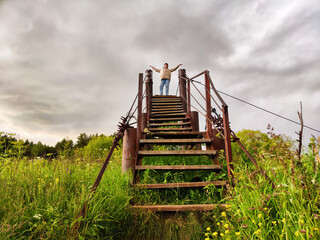 High, old, rusty iron staircase and tiny statue of girl or woman at the top against backdrop of gray, gloomy, thunderous clouds. Concept of an unusual, fantastic journey, surrealism, a strange world