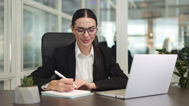 Optimistic successful female sitting at workplace and writing information from digital computer to notebook in office. Focused stylish brunette closed in dark jacket smiling and making notes indoor.