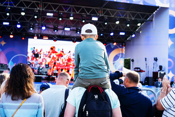 A boy sits on his dad's shoulders during a street concert. A father holds his son in a crowd of people during an open-air festival. Entertainment program of a traveling theater or a musical group - obrazy, fototapety, plakaty