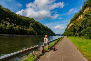 Albert canal between two rocky mountain slopes, senior adult tourist together with her dachshund taking a break, green leafy trees, cyclist in blurred background, sunny day in Kanne, Riemst, Belgium