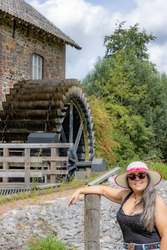 Happy smiling female tourist standing in front of old Eper or Wingbergermolen water mill, hat, sunglasses, tree on blurred background, sunny day in Terpoorten, Epen, South Limburg, Netherlands
