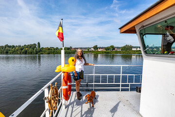 Stern of ferry with woman standing next to her dog crossing Maas river, Belgian flag, yellow plastic duck and a rudder, trees and town in background, sunny day in Eijsden, South Limburg, Netherlands - obrazy, fototapety, plakaty