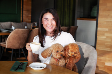Portrait of smiling woman with two small dogs poodles sitting at table in restaurant with cup of coffee. Relationships between people and pets. Beautiful female model in pet friendly cafe