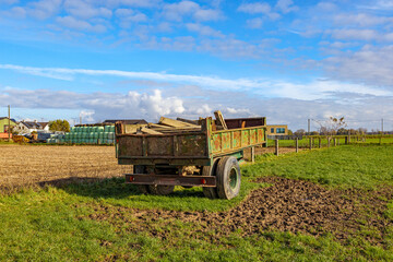 Old trailer car with construction material on agricultural plot, farm with hay bales wrapped in green plastic sheets stacked in background against blue sky, sunny autumn day in Dilsen-Stokkem, Belgium