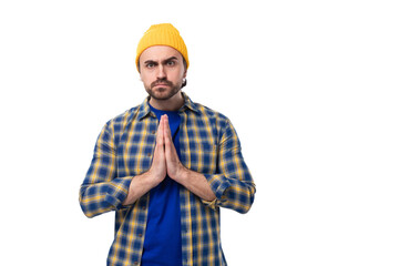 well-groomed young european brunette man with a beard and mustache dressed in a yellow hat and blue shirt put his hands together in hope