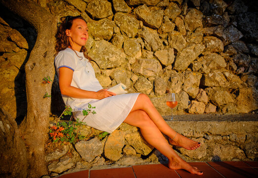 Mature woman sitting with book and wine near stone wall