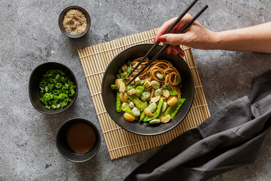 Hand of woman eating bowl of vegan miso udon bowl with tofu, snap peas, broad beans and turnips