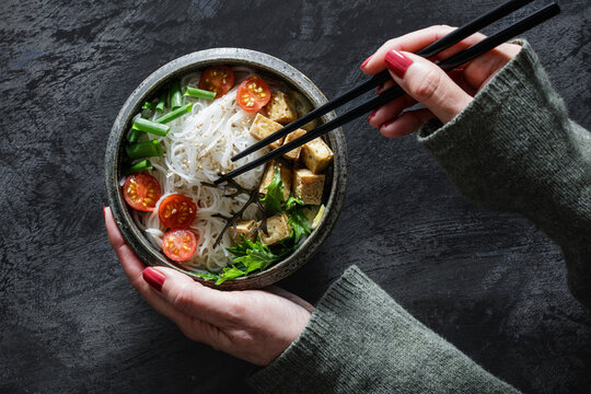 Hands of woman eating bowl of vegan Tomkhakaisoup with tofu, tomatoes, salad, rice noodles, sesame seeds and scallion