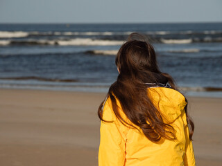 Teenager girl walking on a beach looking at the ocean. Young model in yellow jacket enjoying outdoor activity on fresh air. Selective focus. Cloudy sky. Travel and tourism concept.