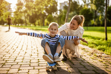 Fototapeta premium Mother having fun with her son in park on sunny day. Boy is ridding skateboard and his mother is pushing him.