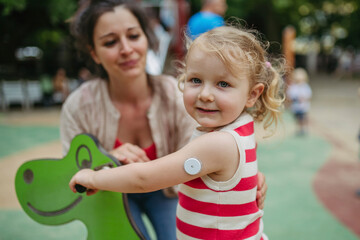 Mother watching little girl with a continuous glucose monitor sitting on dinosaur seesaw on the playground
