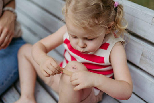 Little girl blowing on injured knee and putting on band aid