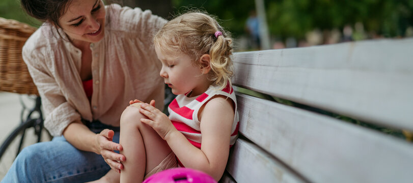 Mother watching little girl blowing on injured knee and putting on band aid