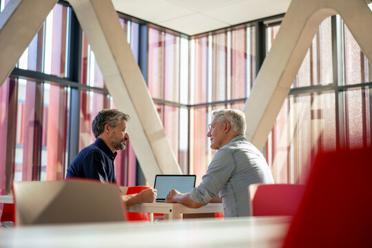 Senior businessmen discussing and sitting with laptop at table in office cafeteria