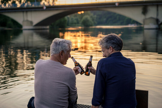 Senior friends toasting beer bottles near water at sunset