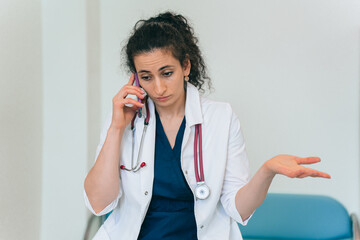 Puzzled brunette female doctor in medical gown with phonendoscope talks by phone with misunderstanding face expression. Tired female doctor exhausted. Medicine, pandemic. - obrazy, fototapety, plakaty