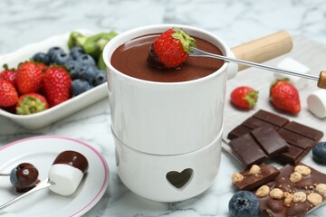Dipping fresh strawberry in fondue pot with melted chocolate at white marble table