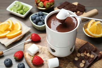 Dipping sweet marshmallow in fondue pot with melted chocolate at wooden table