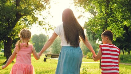 Mother and children run through sunlit park holding hands surrounded by trees
