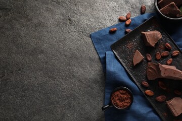 Pieces of tasty milk chocolate, cocoa beans and powder on grey textured table, top view. Space for text