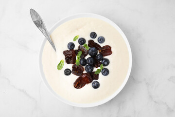 Delicious semolina pudding with blueberries, dates, mint and spoon in bowl on white marble table, top view