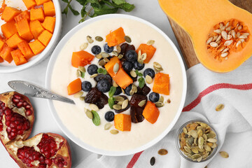 Bowl of delicious semolina pudding with blueberries, dates, pumpkin, spoon and mint on white table, flat lay