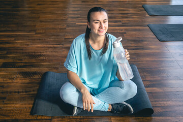 A young woman in blue sportswear sits on a mat and drinks water after a workout.