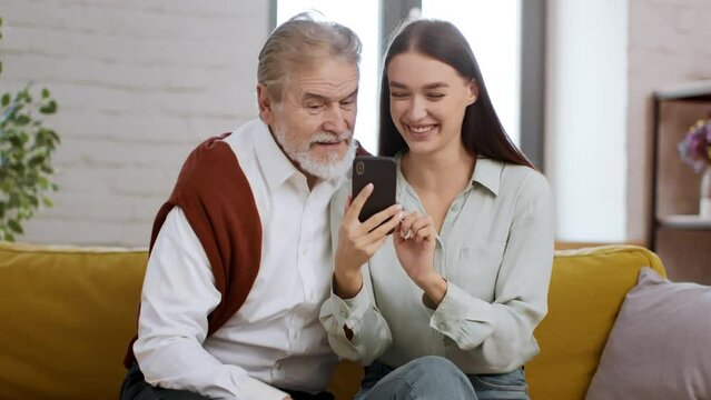 Young happy woman teaching senior grandfather to use mobile application on smartphone, spending time together at home