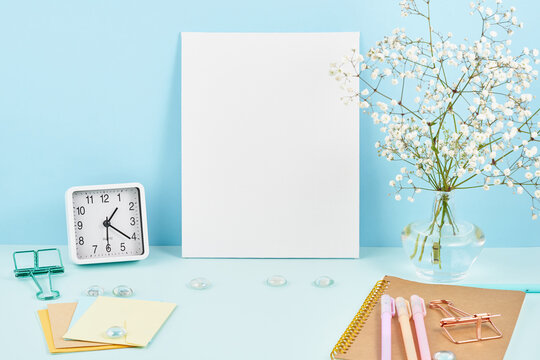Mockup with blank white frame on blue table against blue wall, alarm, flower in vaze.