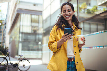 Woman texting and drinking coffee outdoors. Young smiling brunette woman holding a smartphone and a...