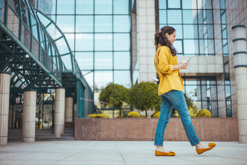 Woman texting and drinking coffee outdoors. Young smiling brunette woman holding a smartphone and a cup of coffee looking at device screen against the background of an office building.