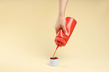 Woman squeezing tasty ketchup from bottle into bowl on beige background, closeup