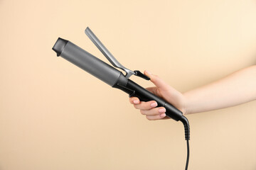 Hair styling appliance. Woman holding curling iron on beige background, closeup