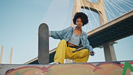 Bright girl posing skateboard at urban bridge. African skater sitting street