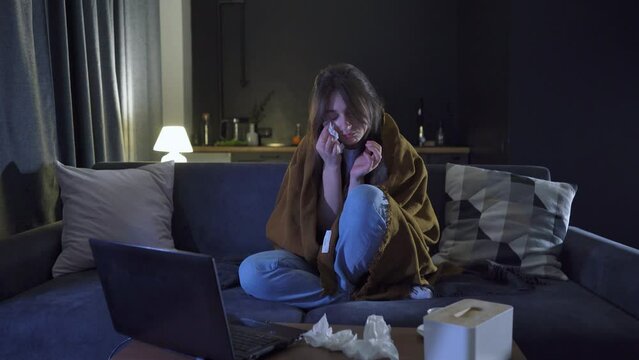 A sad woman behind a laptop screen cries while watching a video and wipes her tears with a napkin. The girl is depressed. Stress concept. A sad Caucasian girl is watching a melodrama movie at home.