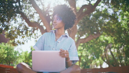 Curly teenager sitting laptop at sunlight looking around. African girl relaxing 