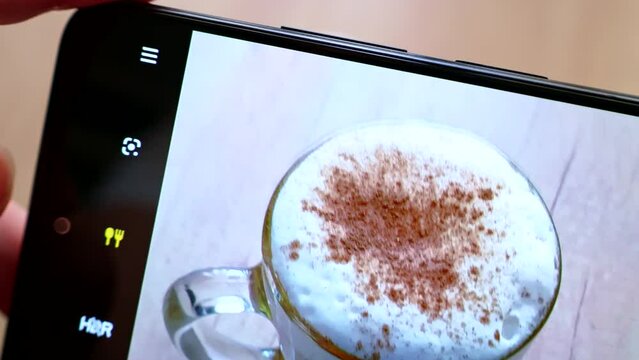 Woman hands taking photos by modern smartphone of hot cappuccino coffee cup on wooden table. Making pictures for social network. Phone screen close up