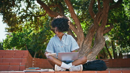 Young freelancer writing notes in notepad sitting park with laptop. Girl webinar