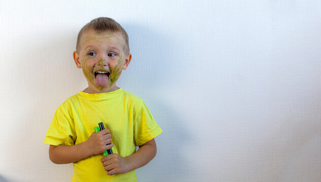 A 3-year-old boy painted his face with a marker. The boy is standing against a white wall, holding markers in his hands, smiling and showing his tongue. Banner with space for text