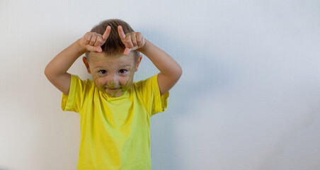 A 3-year-old boy painted his face with a marker. The boy stands at the white wall and makes horns. Banner with space for text