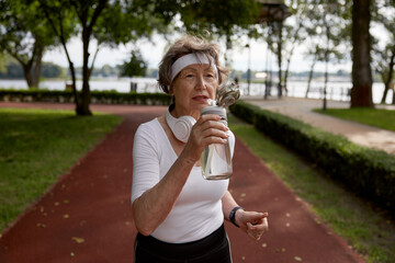 Fototapeta premium Elderly woman drinking water taking short break during running exercise