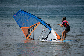 Naklejka premium young sporty girl on the windsurf board raising the sail pulling the rope