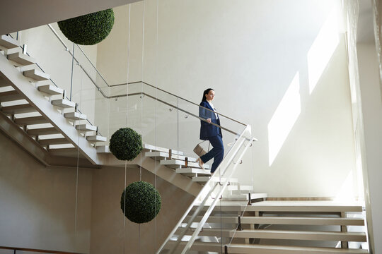 Wide angle shot of Caucasian woman in business suit carrying purse going downstairs holding on handrail in modern office building, copy space