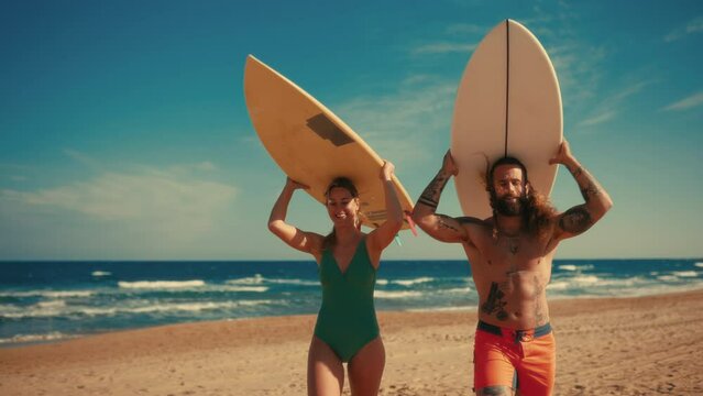 A couple of surfers on the beach. Caucasian woman in a swimsuit and a bearded buff man with tattoos in swimming trunks with surfboards near the sea. Sports and active recreation.