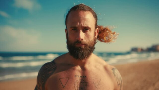 A muscular, athletic bearded man with long hair and tattoos standing on a sandy beach on the seashore in sunny weather. Tourism and rest.