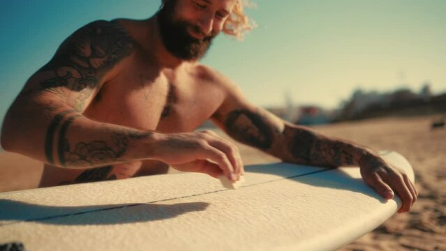 Caucasian beefy bearded man with tattoos in swimming trunks is waxing a surfboard near the sea. Sports and active recreation.