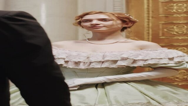 Vertical medium slowmo of young beautiful Caucasian woman wearing charming Regency dress looking at her male companion while making elegant curtsy in luxury ballroom