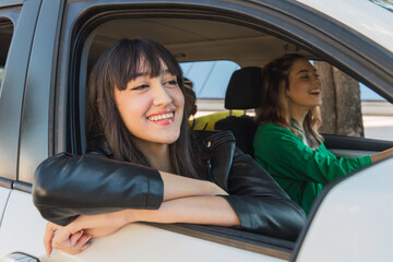 Cheerful ethnic woman sitting in parked white car near tree trunk