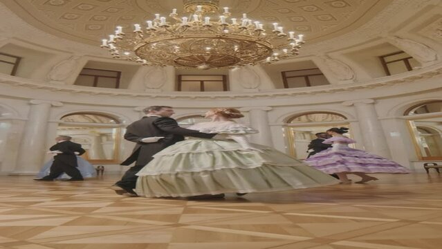 Vertical full length slowmo of people in fancy medieval outfits ball dancing in pairs in classic ballroom with gold crystal chandelier on high ceiling