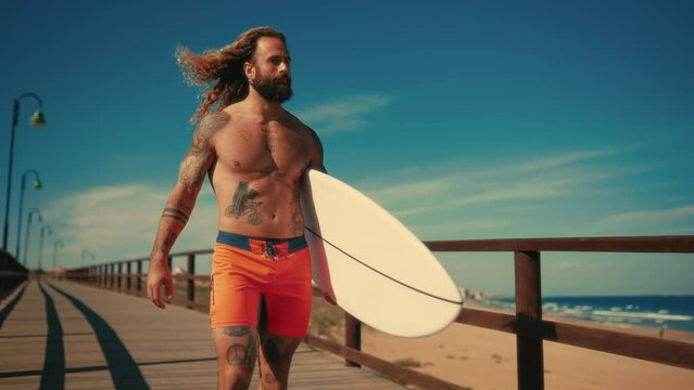 Caucasian beefy bearded man with tattoos in swimming trunks holding a surfboard walks along a wooden bridge to the sea. Sports and active recreation.